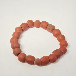 Könnte beinhalten: Ein kreisförmiges Armband aus rotbraunen, unregelmäßig geformten Perlen. Die Perlen variieren in Größe und Textur, wobei einige glatter erscheinen als andere. Das Armband ist vor einem schlichten weißen Hintergrund platziert.