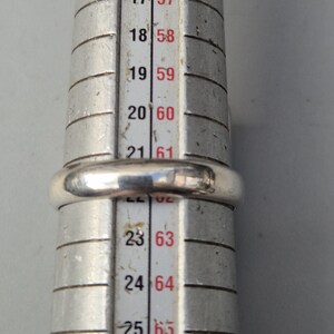 May include: A silver ring is placed on a ring sizer. The ring is a simple band design. The ring sizer has measurements in both US and European sizes. The ring appears to be a size 20 (US size 9.5) or 60 (European size 19.1).
