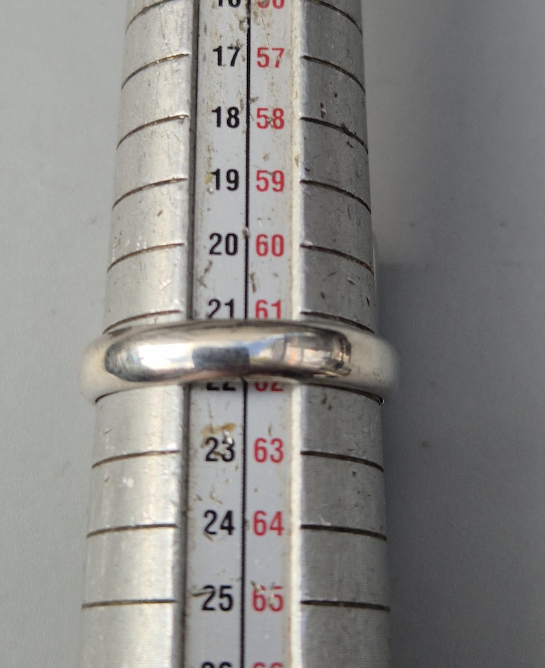 May include: A silver ring is placed on a ring sizer. The ring is a simple band design. The ring sizer has measurements in both US and European sizes. The ring appears to be a size 20 (US size 9.5) or 60 (European size 19.1).