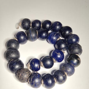 May include: A strand of dark blue glass beads with a variety of textures and shapes. Some beads are smooth and glossy, while others have a rough, aged appearance. The beads are strung together with silver-colored metal findings, creating a necklace.