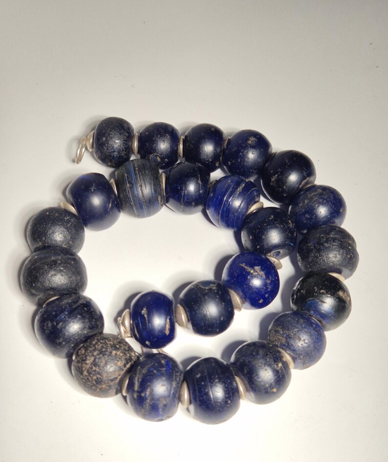May include: A strand of dark blue glass beads with a variety of textures and shapes. Some beads are smooth and glossy, while others have a rough, aged appearance. The beads are strung together with silver-colored metal findings, creating a necklace.