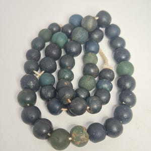Könnte beinhalten: Eine Halskette aus runden Perlen in den Farbtönen Dunkelblau, Grün und Türkis. Die Perlen sind mit einer naturfarbenen Kordel aufgefädelt. Die Perlen haben ein leicht verwittertes Aussehen.