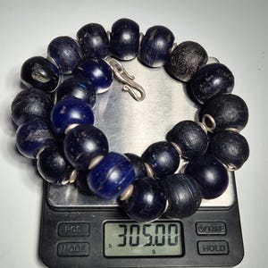 May include: A dark blue beaded necklace with a silver clasp, resting on a digital scale. The scale displays a weight of 305.00 grams. The beads are large and appear to be made of glass or a similar material.