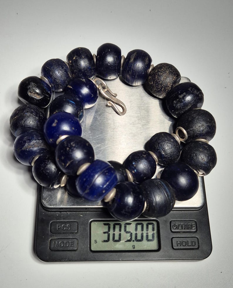 May include: A dark blue beaded necklace with a silver clasp, resting on a digital scale. The scale displays a weight of 305.00 grams. The beads are large and appear to be made of glass or a similar material.