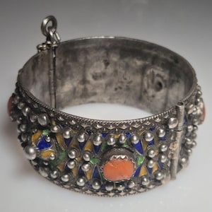 May include: A silver-toned hinged bracelet with colorful enamel and coral-colored stone accents. The bracelet features a detailed design with small, round embellishments and geometric patterns in blue, yellow, and green. The clasp is visible on the side.