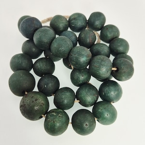 Könnte beinhalten: Eine Halskette aus dunkelgrünen, runden Perlen, die auf einer naturfarbenen Kordel aufgefädelt sind. Die Perlen haben ein leicht verwittertes Aussehen mit Farb- und Texturvariationen. Die Halskette wird vor weißem Hintergrund präsentiert.