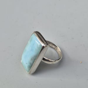 May include: A rectangular silver ring featuring a large, light blue gemstone. The gemstone has a marbled appearance. The ring's band is smooth and polished. The ring is set against a neutral background.