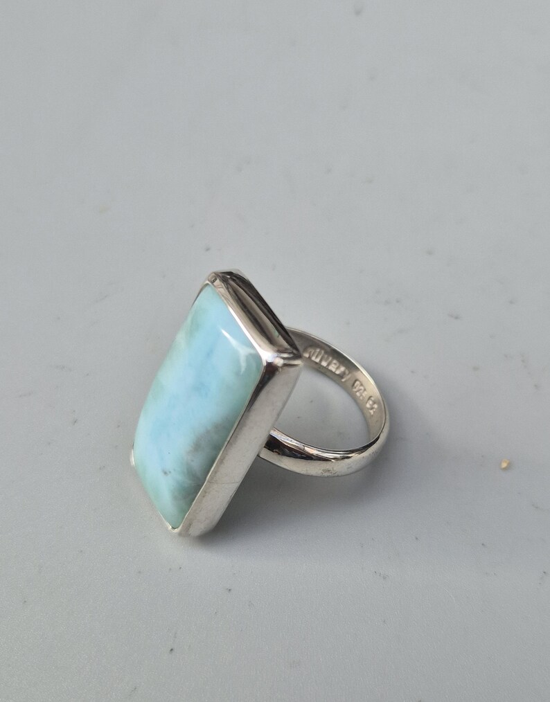 May include: A rectangular silver ring featuring a large, light blue gemstone. The gemstone has a marbled appearance. The ring's band is smooth and polished. The ring is set against a neutral background.