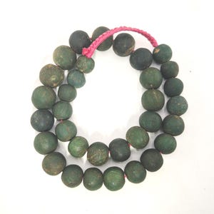 Könnte beinhalten: Eine Halskette mit dunkelgrünen, runden Perlen, die auf einer rosa Schnur aufgefädelt sind. Die Perlen haben eine strukturierte Oberfläche und variieren leicht in der Größe. Die Halskette wird vor einem weißen Hintergrund gezeigt.