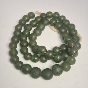 Könnte beinhalten: Eine Halskette aus dunkelgrünen, runden Perlen, die auf einer Naturfaserschnur aufgefädelt sind. Die Perlen variieren leicht in der Größe und haben eine matte Oberfläche. Die Halskette wird vor weißem Hintergrund präsentiert.