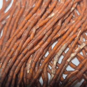May include: Close-up of a collection of orange-brown beads. The beads are cylindrical, with a slightly irregular shape and a matte finish. They are strung together, creating a tangled, textured appearance. The color varies slightly, with some beads appearing darker than others.