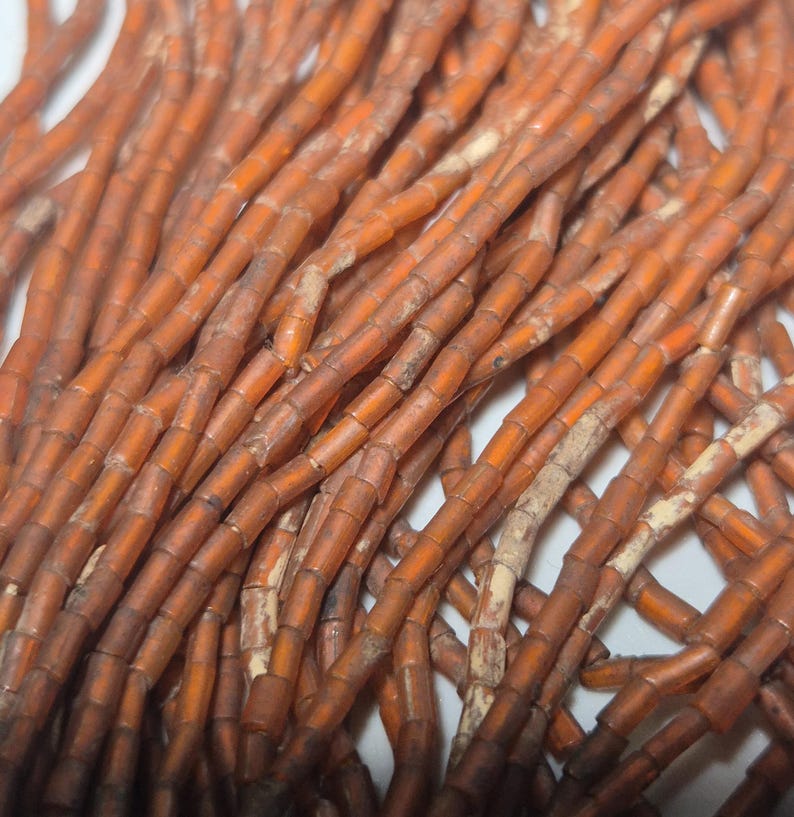 May include: Close-up of a collection of orange-brown beads. The beads are cylindrical, with a slightly irregular shape and a matte finish. They are strung together, creating a tangled, textured appearance. The color varies slightly, with some beads appearing darker than others.