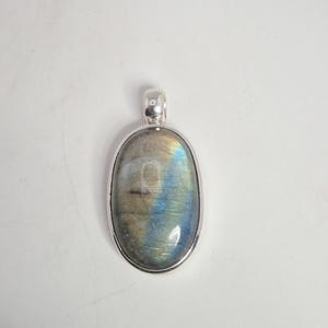 May include: Oval labradorite pendant with a silver-colored bezel. The gemstone displays iridescent blue, green, and yellow hues. The pendant has a loop at the top for a chain. A beautiful piece of jewelry.