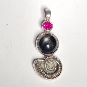 Könnte beinhalten: Silberanhänger mit einem rosa Edelstein, einer schwarzen Kugel und einem versteinerten Ammoniten. Der Anhänger hat oben eine Schlaufe für eine Kette. Das Ammonitenfossil ist hellgrau.