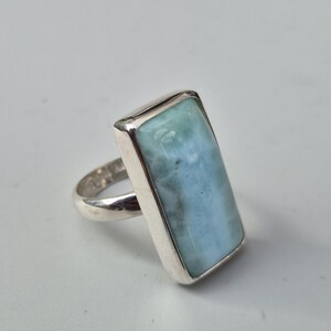 May include: A rectangular silver ring featuring a large, light blue gemstone with a marbled appearance. The gemstone is set in a polished silver band, creating a simple yet elegant design. The ring is shown on a white background.