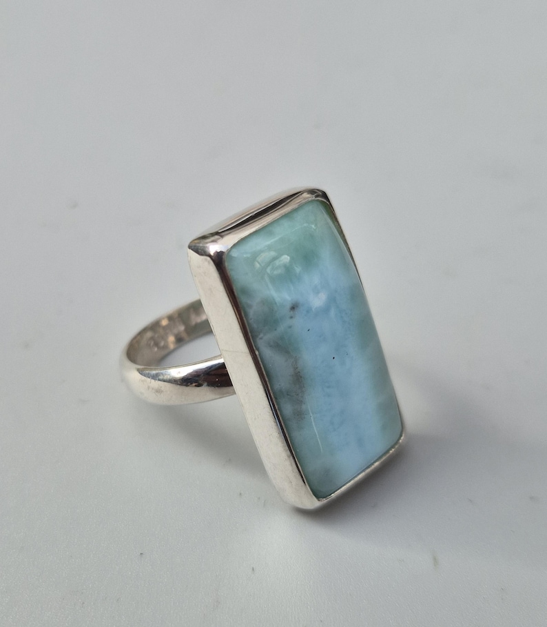 May include: A rectangular silver ring featuring a large, light blue gemstone with a marbled appearance. The gemstone is set in a polished silver band, creating a simple yet elegant design. The ring is shown on a white background.
