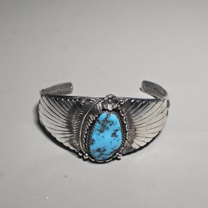 Peut inclure: Un bracelet manchette en argent avec une grosse pierre ovale turquoise au centre. Le bracelet présente des motifs détaillés en forme d'ailes de chaque côté de la pierre et un motif floral. La turquoise est bleu vif avec une matrice sombre.
