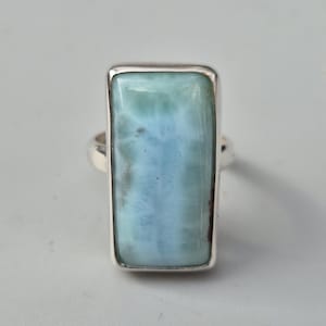 May include: A rectangular ring featuring a large, light blue larimar stone set in a silver band. The stone has a marbled appearance with shades of blue and white, and is framed by a polished silver bezel.
