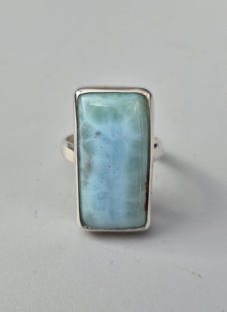 May include: A rectangular ring featuring a large, light blue larimar stone set in a silver band. The stone has a marbled appearance with shades of blue and white, and is framed by a polished silver bezel.