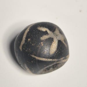 May include: A dark, rounded bead with a black base and white, abstract markings. The bead has a hole running through the center, suggesting it is for stringing. The markings appear to be a stylized design.