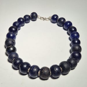 May include: A necklace featuring large, dark blue beads with a silver clasp. The beads vary in color and texture, with some appearing almost black. The necklace is displayed against a plain white background.