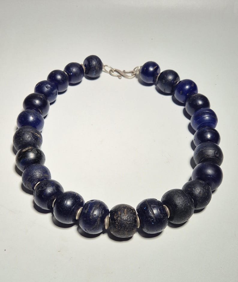 May include: A necklace featuring large, dark blue beads with a silver clasp. The beads vary in color and texture, with some appearing almost black. The necklace is displayed against a plain white background.