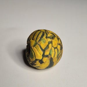 Könnte beinhalten: Eine runde, gelbe Perle mit einem Mosaikmuster aus schwarzen und grünen Formen. Die Perle hat eine leicht unregelmäßige Form und scheint aus Stein oder Glas zu bestehen. Es ist ein dekorativer Gegenstand.