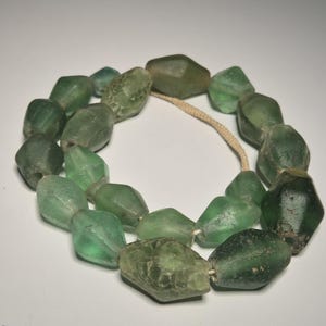 Könnte beinhalten: Eine Halskette aus grünen Glasperlen. Die Perlen sind unregelmäßig geformt und auf einer beigefarbenen Schnur aufgefädelt. Das Glas hat eine leicht durchscheinende Qualität, mit Farbvariationen von hell- bis dunkelgrün.
