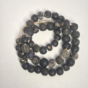 Könnte beinhalten: Eine Halskette oder ein Armband mit dunklen, runden Perlen mit einem verwitterten, rustikalen Aussehen. Die Perlen sind überwiegend schwarz, einige weisen hellere, strukturierte Stellen auf. Sie sind auf einer naturfarbenen Kordel aufgefädelt.