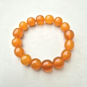 May include: A bracelet featuring a strand of polished, amber-colored beads. The beads are round and have a glossy finish, strung together to form a circular shape. The warm, orange-yellow hues of the beads create a vibrant and eye-catching accessory.
