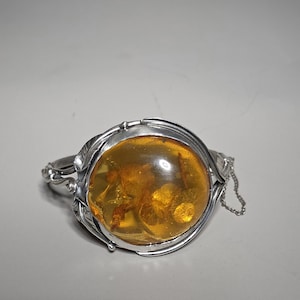 Könnte beinhalten: Ein silberfarbenes Armband mit einem großen, ovalen Bernstein. Der Bernstein hat eine warme, goldene Farbe mit sichtbaren Einschlüssen. Das Armband hat ein verziertes, florales Design um den Bernstein und einen zarten Kettenverschluss.