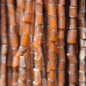 May include: Close-up of strands of amber-colored beads. The beads are cylindrical with rounded edges, creating a textured surface. The color varies from light to dark amber, with some beads appearing more opaque than others. The beads are strung together, forming vertical lines.