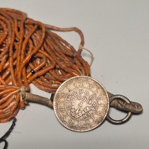 May include: An antique necklace featuring a large silver coin pendant with intricate engravings and a ring clasp. The necklace includes strands of small, orange-brown beads. The coin has text and a central design.