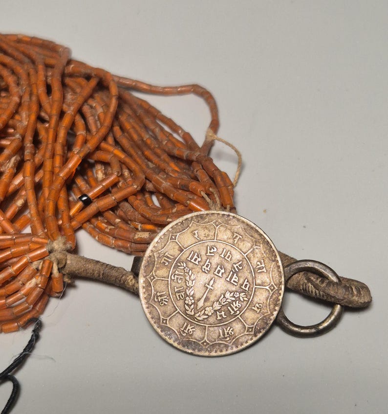 May include: An antique necklace featuring a large silver coin pendant with intricate engravings and a ring clasp. The necklace includes strands of small, orange-brown beads. The coin has text and a central design.
