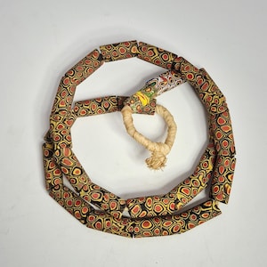 Può includere: Una collana composta da perline cilindriche con un motivo ripetuto di cerchi rossi e gialli su sfondo nero. La collana è collegata da un cordoncino beige intrecciato.