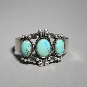 May include: A silver cuff bracelet with three oval turquoise stones. The stones are set in decorative frames with bows and small silver beads. The bracelet has a polished finish and a classic Southwestern design.