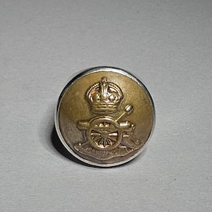 Könnte beinhalten: Ein runder, antiker Messingknopf mit silbernem Rand. Der Knopf zeigt ein detailliertes Emblem einer Krone über einer Kanone. Die Kanone hat ein Rad mit Speichen und steht auf einem dekorativen Sockel. Wahrscheinlich ein militärischer oder historischer Artikel.