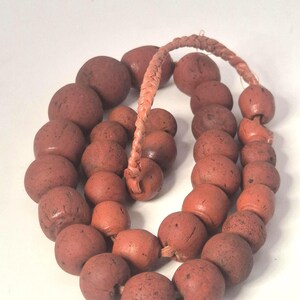 Könnte beinhalten: Eine Halskette aus großen, runden, rotbraunen Perlen. Die Perlen sind auf einer geflochtenen, hellbraunen Kordel aufgefädelt. Die Perlen haben eine leicht raue Textur und sind unterschiedlich groß. Die Halskette wird auf einer weißen Oberfläche präsentiert.