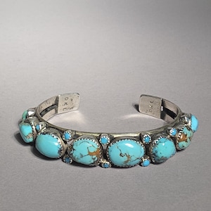 May include: A silver cuff bracelet with oval turquoise stones. The bracelet features a row of large turquoise stones with smaller turquoise stones in between. The bracelet is open at the ends and has a simple design.