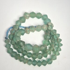 Könnte beinhalten: Kette aus hellgrünen Glasperlen. Die Perlen sind facettiert und unregelmäßig geformt, auf einer blauen Schnur aufgefädelt. Die Perlen variieren in Größe und Form und wirken antik. Die Perlen sind kreisförmig angeordnet.