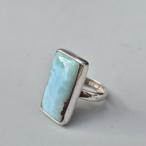 May include: A rectangular silver ring featuring a large, light blue gemstone with brown inclusions. The gemstone is set in a polished silver band. The ring is a statement piece, suitable for everyday wear or special occasions.