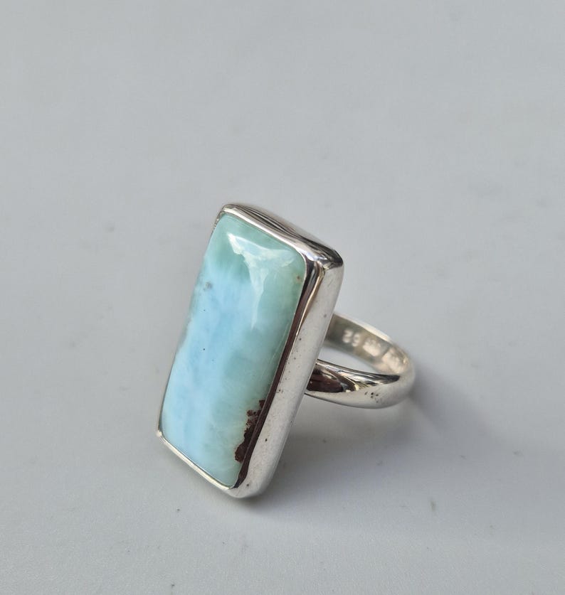 May include: A rectangular silver ring featuring a large, light blue gemstone with brown inclusions. The gemstone is set in a polished silver band. The ring is a statement piece, suitable for everyday wear or special occasions.