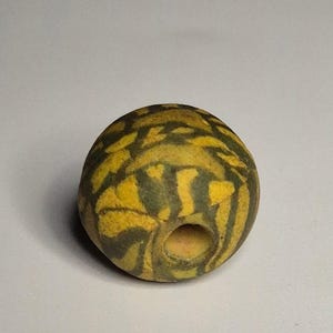 Könnte beinhalten: Eine runde, gelbe Perle mit einem dunkelgrünen, gestreiften Muster. Die Perle hat ein Loch in der Mitte. Die Perle ist wahrscheinlich aus Stein oder Glas und eignet sich für die Schmuckherstellung oder andere Bastelarbeiten.