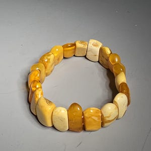 May include: A bracelet made of amber-colored stones. The stones vary in shape and shade, ranging from light cream to deep honey hues. The bracelet is strung together, forming a circular shape. The stones are polished and have a smooth texture.