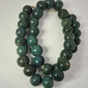 Könnte beinhalten: Zwei Stränge dunkelgrüner, runder Perlen, vermutlich aus Stein oder Glas, sind auf einer hellen Schnur aufgereiht. Die Perlen variieren leicht in der Farbe, einige erscheinen in einem tieferen, fast schwarzen Grün, andere in einem helleren Türkisgrün.