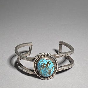 May include: Silver cuff bracelet with a large oval turquoise stone. The bracelet has a double band design with starburst and dot details. The turquoise stone is set in a beaded bezel. The bracelet is approximately 15 cm in diameter.