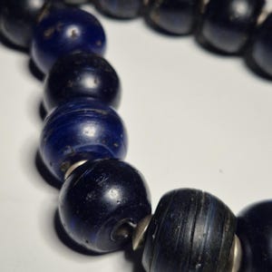 May include: A close-up of a necklace featuring large, dark blue beads. The beads are round and have a slightly textured appearance. Silver-colored metal spacers are between each bead. The necklace is on a white background.