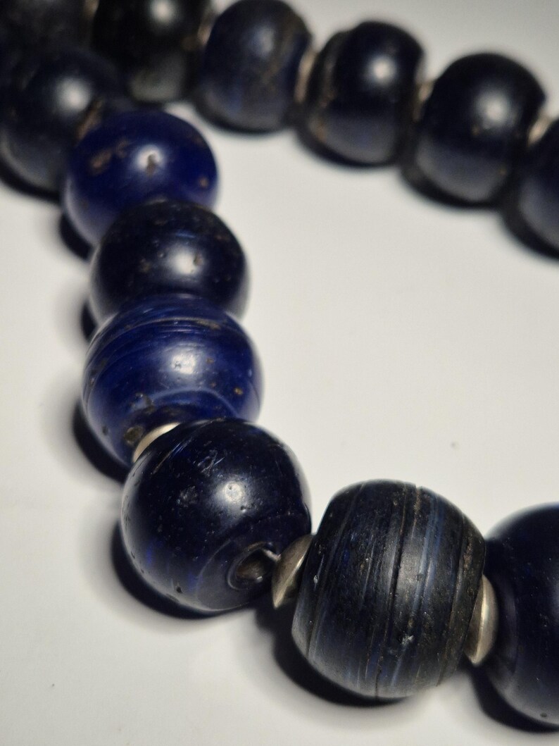 May include: A close-up of a necklace featuring large, dark blue beads. The beads are round and have a slightly textured appearance. Silver-colored metal spacers are between each bead. The necklace is on a white background.