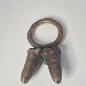 Könnte beinhalten: Ein bronzefarbener Ring im Vintage-Stil mit zwei nach unten zeigenden, zahnförmigen Anhängern. Der Ring ist kreisförmig und die Anhänger haben eine strukturierte Oberfläche. Das Stück hat ein gealtertes Aussehen.