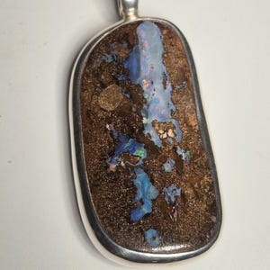 Könnte beinhalten: Ein großer, polierter Opalanhänger mit silberfarbener Öse. Der Opal hat eine dunkelbraune Matrix mit leuchtenden blauen, grünen und roten Blitzen. Der Anhänger hat eine rechteckige Form mit abgerundeten Kanten.